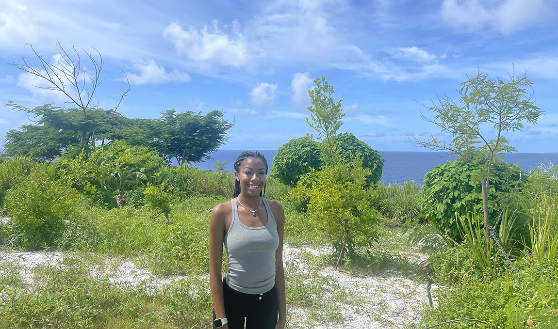 A Black woman with brown hair and athletic attire standing in a grassy field neutrally posing for a picture